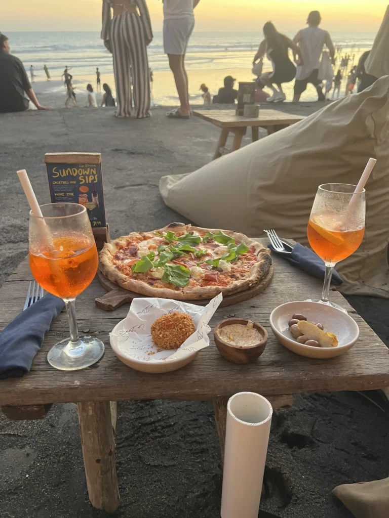 Dinner setup on the beach in Canggu, Bali, with two Aperol Spritz cocktails, a pizza, and appetizers on a wooden table during sunset, people relaxing in the background by the ocean.