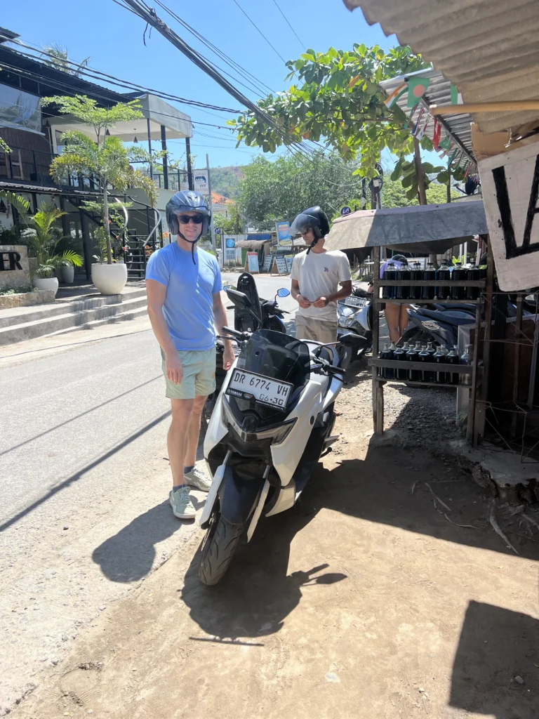 Two people wearing helmets standing next to a white scooter on a sunny street in Southeast Asia, surrounded by shops and parked motorbikes.