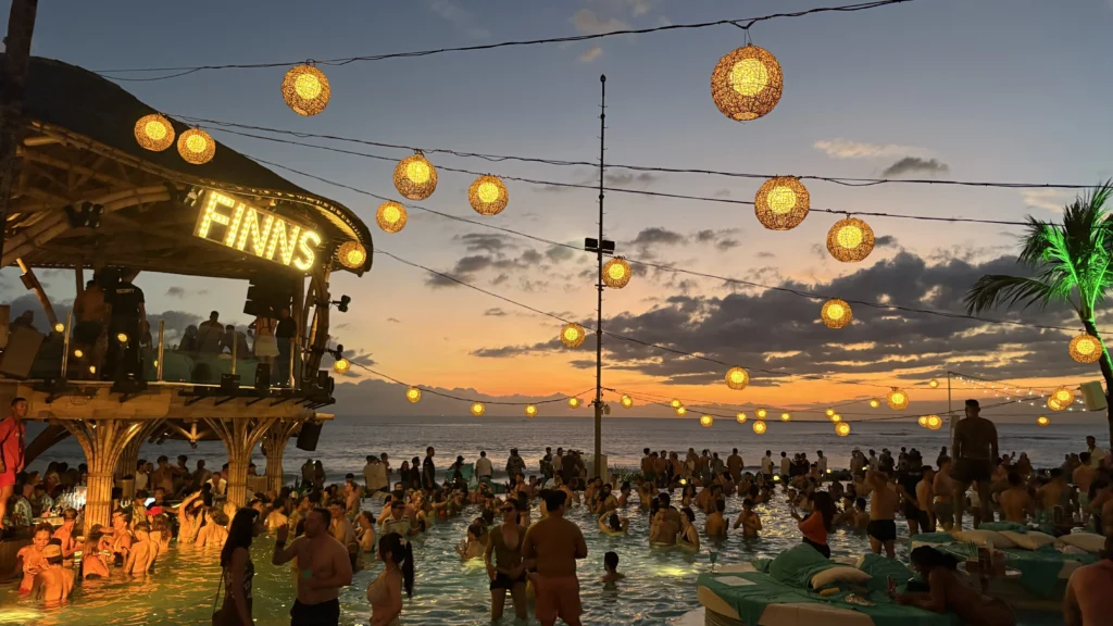 Crowded pool party at Finns Beach Club in Canggu, Bali, with glowing lanterns above and a colorful sunset over the ocean in the background.
