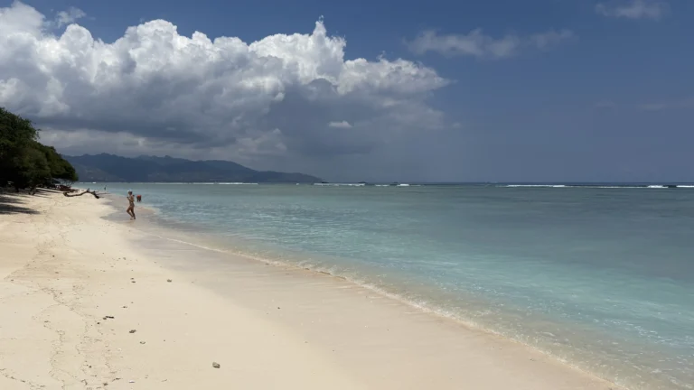Peaceful tropical beach on the Gili Island with white sand, turquoise water, and a few people walking along the shoreline under a partly cloudy sky.