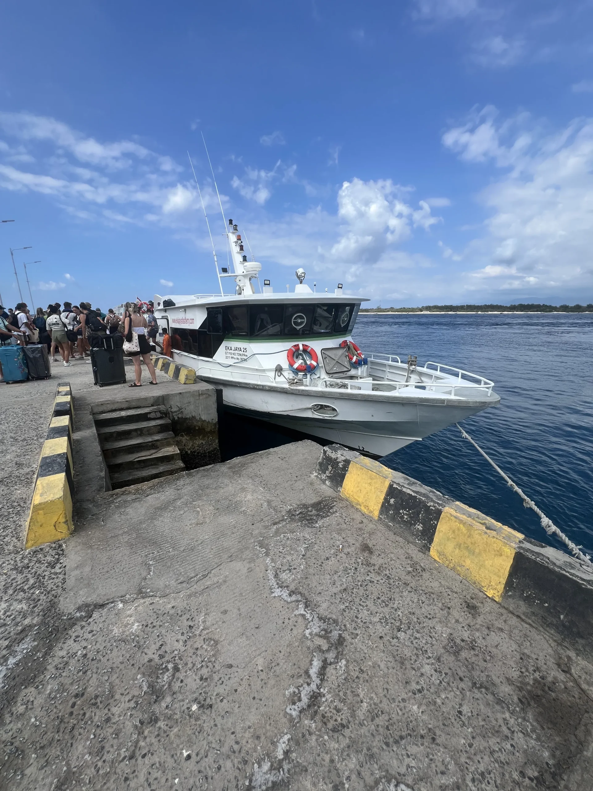 Fast boat ferry with travelers boarding and clear blue ocean water under a bright sky with scattered clouds.