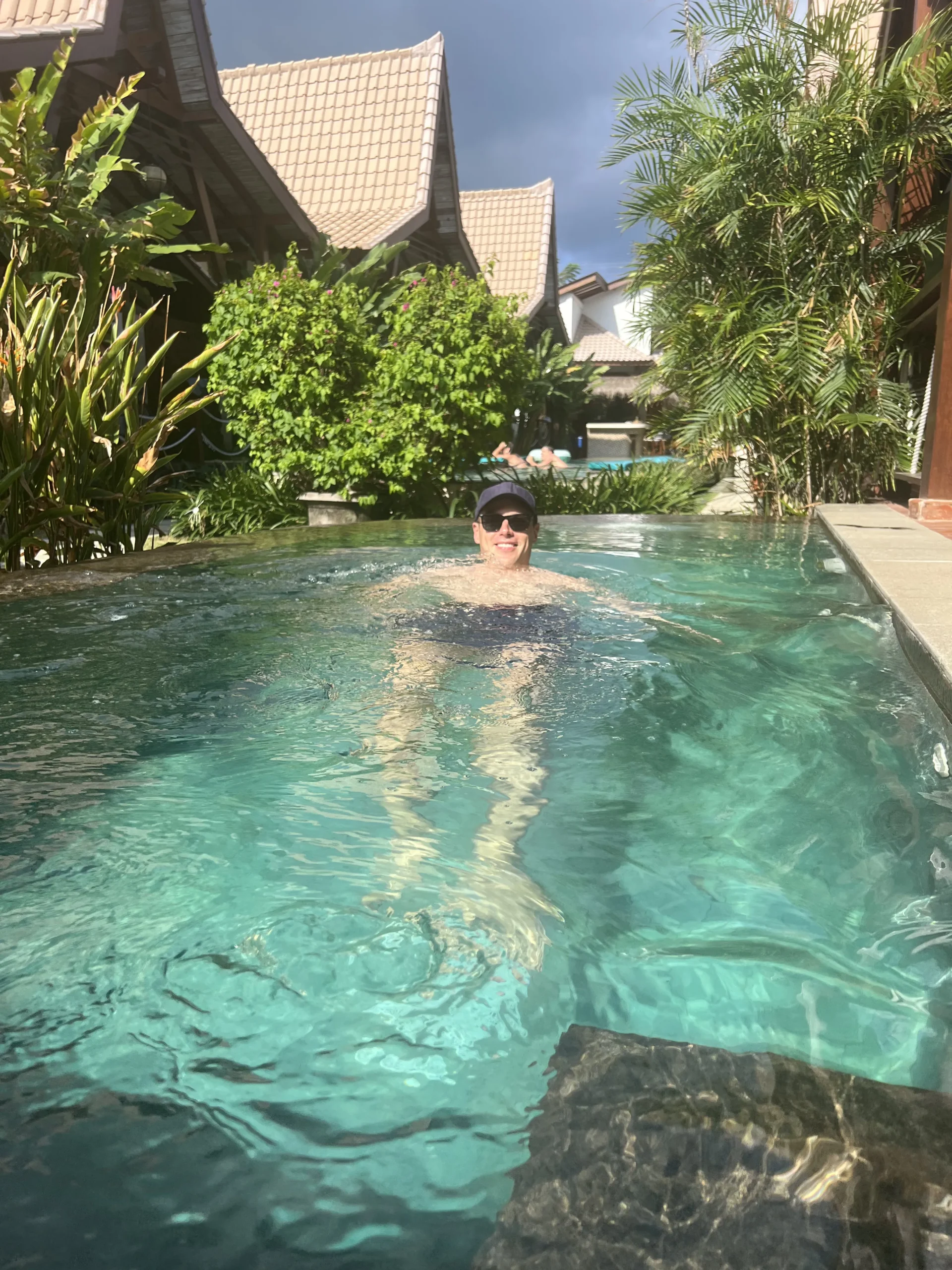 Man swimming in a clear turquoise pool at a tropical hotel on the Gili Islands, surrounded by palm trees and villas under a bright sunny sky.
