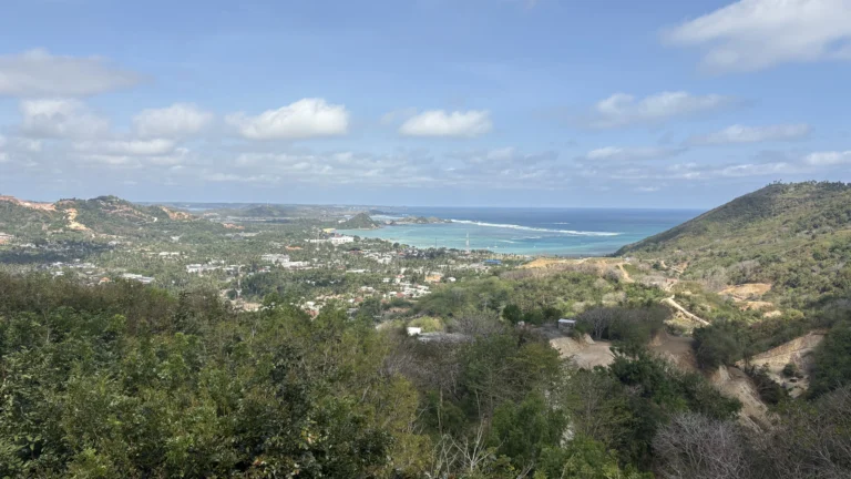 Scenic panoramic view of Kuta Lombok, Indonesia, showing green hills, a winding coastline, and turquoise ocean water under a bright blue sky.