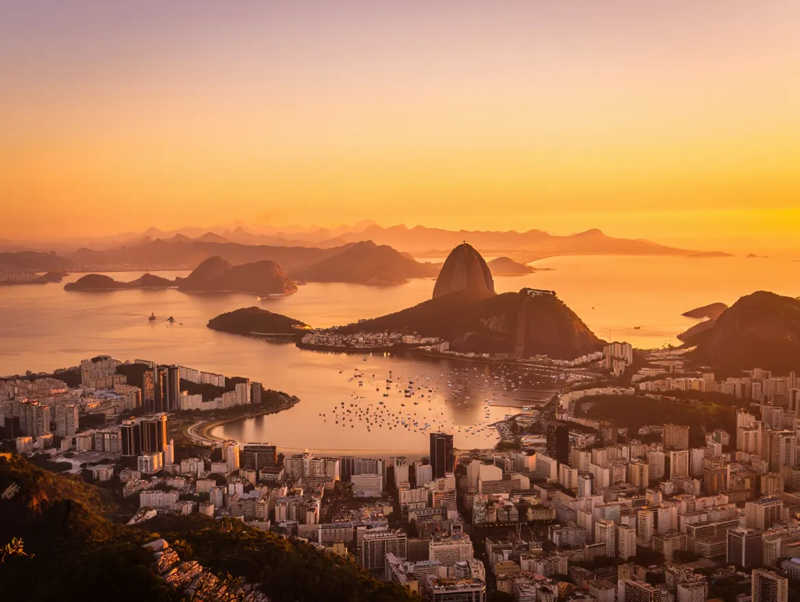 A stunning sunset panorama of Rio de Janeiro, Brazil, from Sugarloaf Mountain, featuring the iconic Christ the Redeemer statue overlooking the city and Guanabara Bay as the sky glows with golden light.