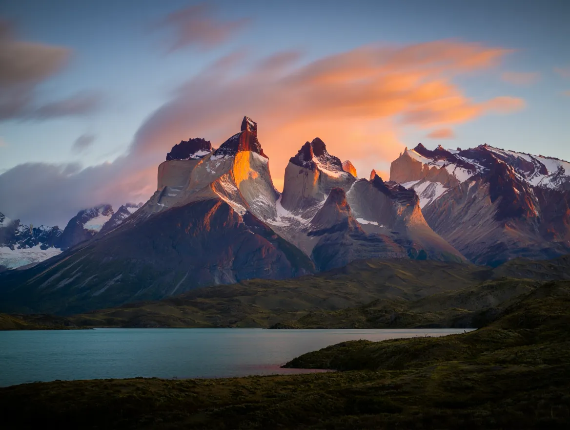The iconic granite peaks of Torres del Paine in Chilean Patagonia at sunrise, glowing with pink and golden light and perfectly reflected in the calm, turquoise water of a glacial lake.