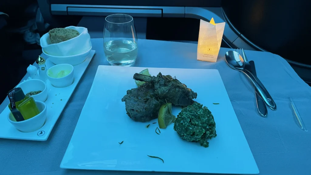 Turkish Airlines Business Class dinner, grilled lamb chops with spinach, bread basket, olive oil, and a glass on the tray table under blue cabin lighting.
