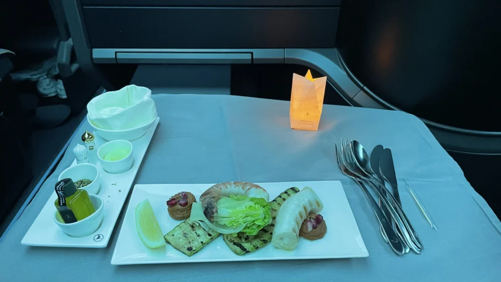Turkish Airlines Business Class appetizer plate with prawn, grilled zucchini, white cheese roll, pomegranate dip, lemon wedge, olive oil set, and bread basket under blue cabin lighting.