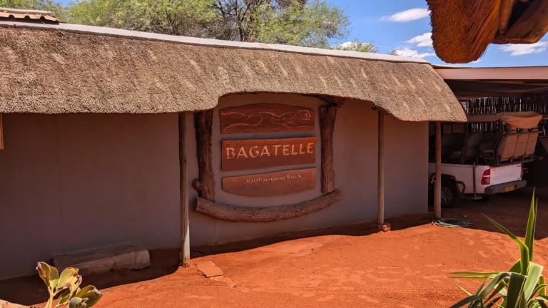 Entrance sign for Bagatelle Lodge mounted on a thatched wall above red desert sand.
