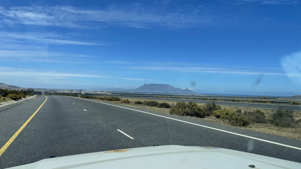 Highway leading toward Cape Town with a clear view of Table Mountain under a bright blue sky, seen from inside a moving vehicle.