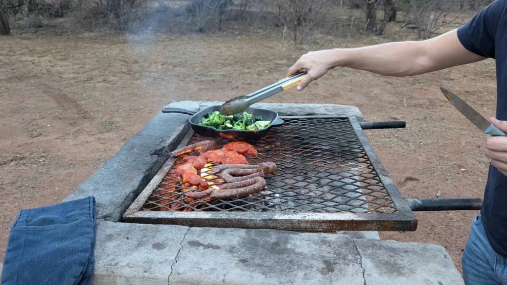 Person grilling outdoors on a rustic stone barbecue, cooking sausages, marinated meat, and broccoli in a cast-iron pan over open flames in a dry bush setting.