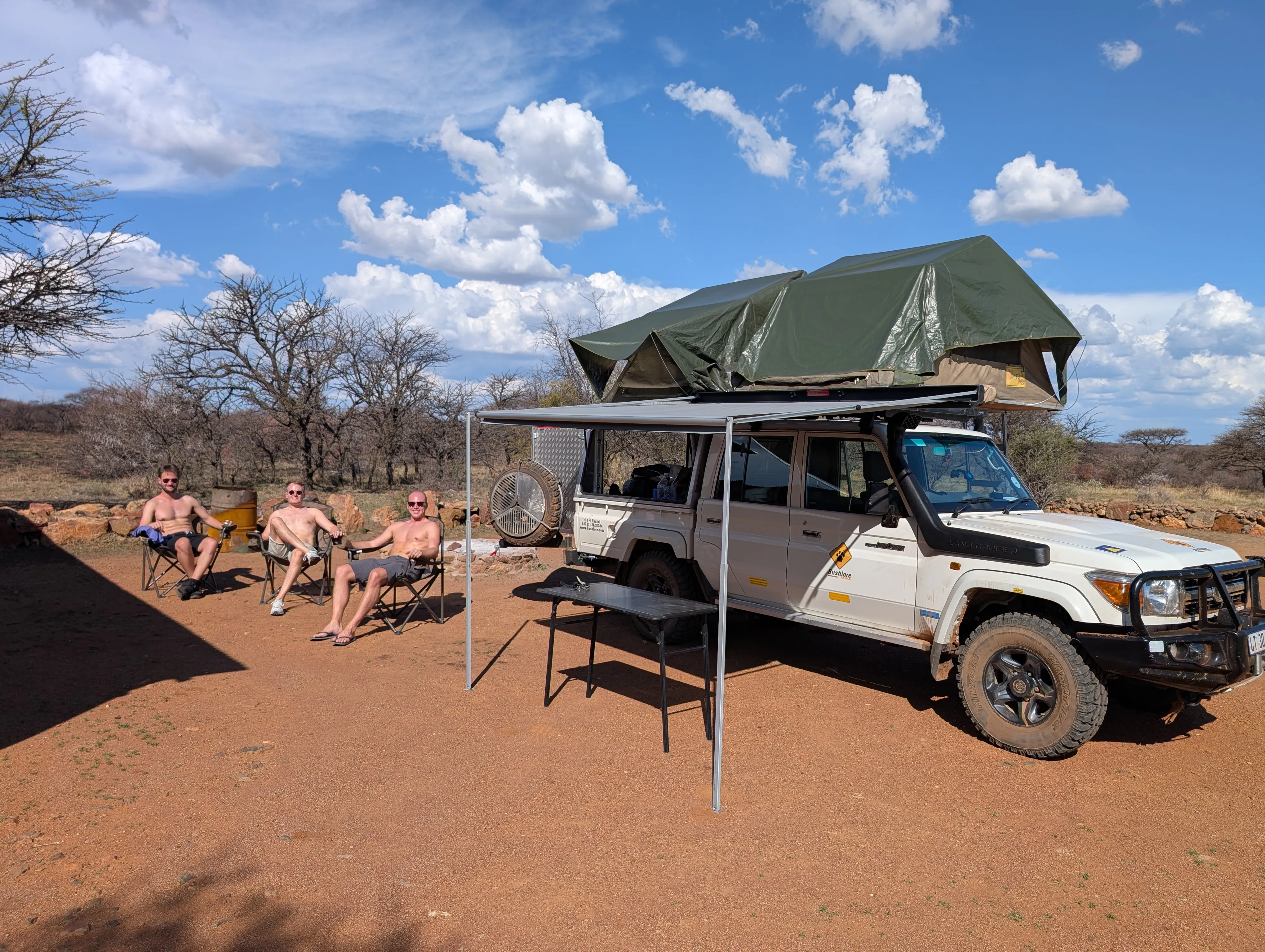 Group relaxing beside an off-road vehicle in Botsalano Game Reserve with a rooftop tent and awning set up at a sunny campsite on red soil with blue skies and scattered clouds.