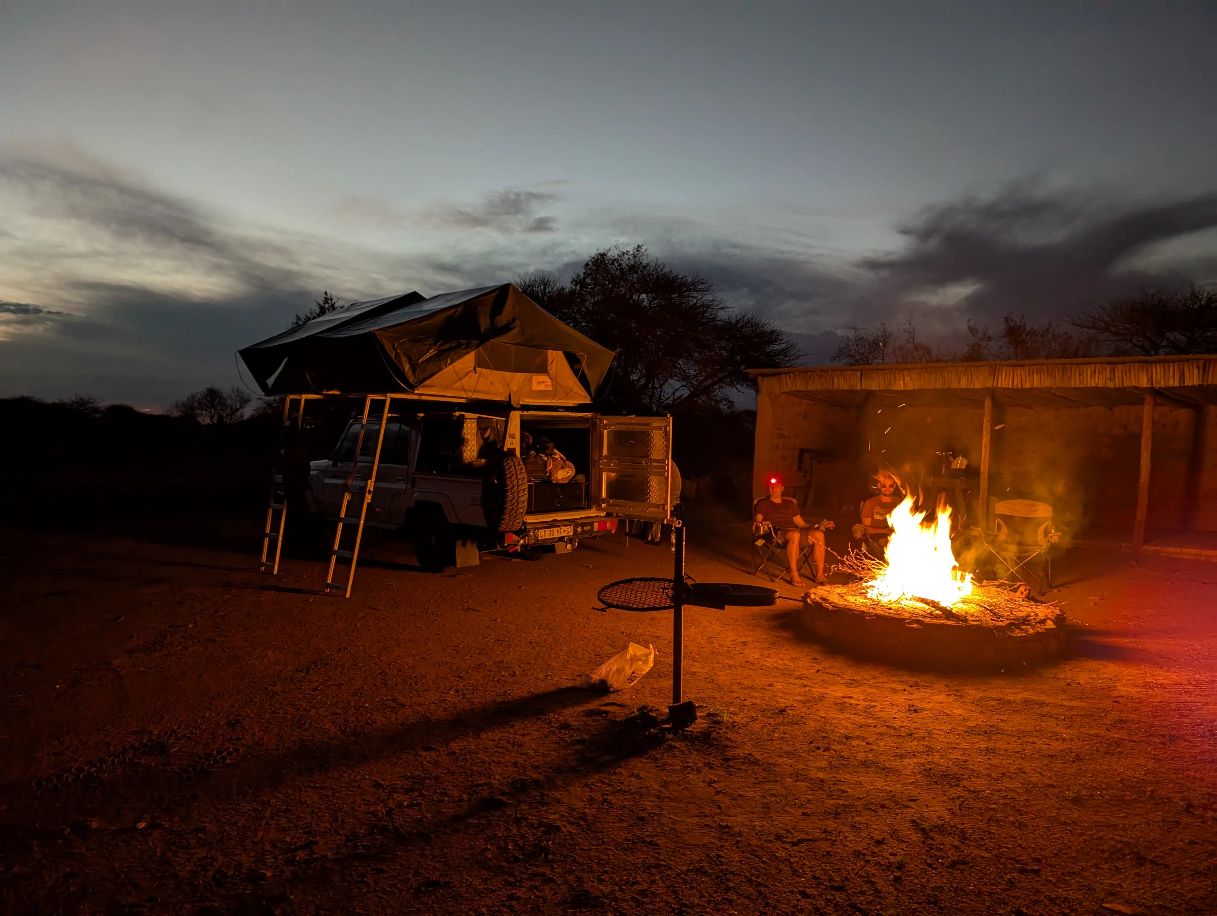 Nighttime campsite scene with a rooftop tent vehicle and a glowing campfire illuminating the area under a darkening sky.