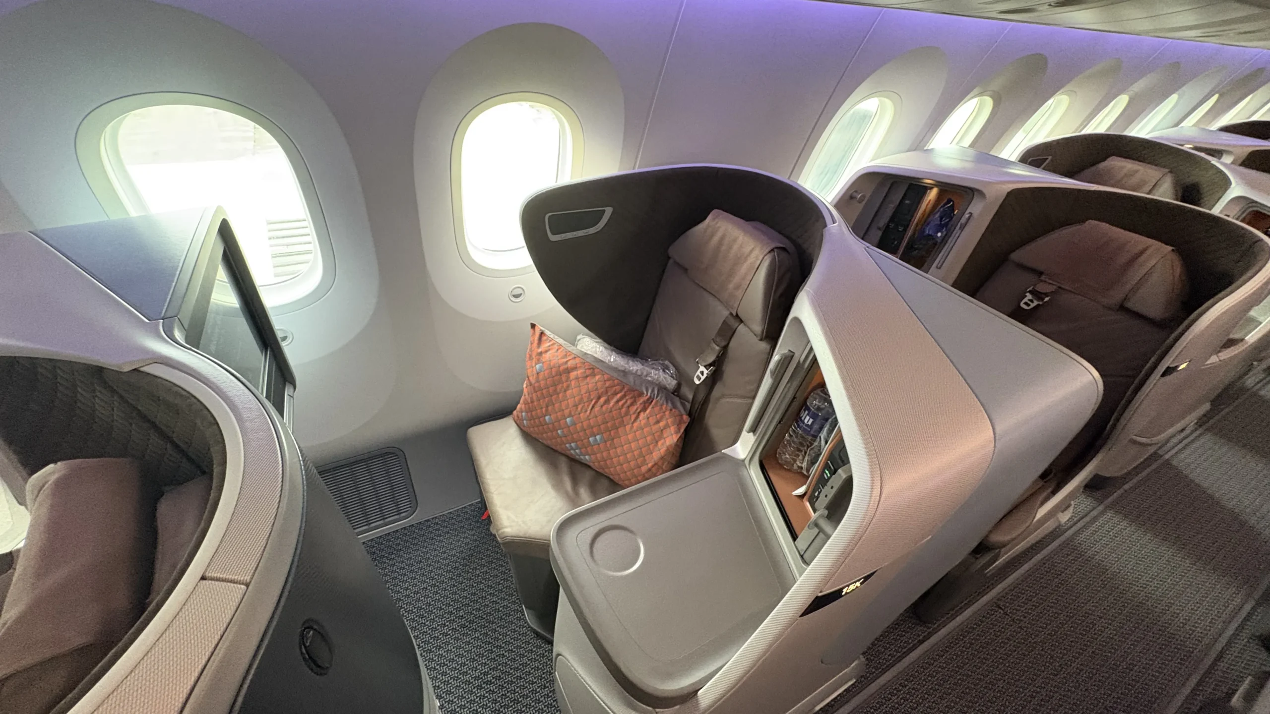 Modern business-class seat on a wide-body aircraft with a private shell design, large side console, lie-flat seating, and soft mood lighting, positioned next to multiple large windows.