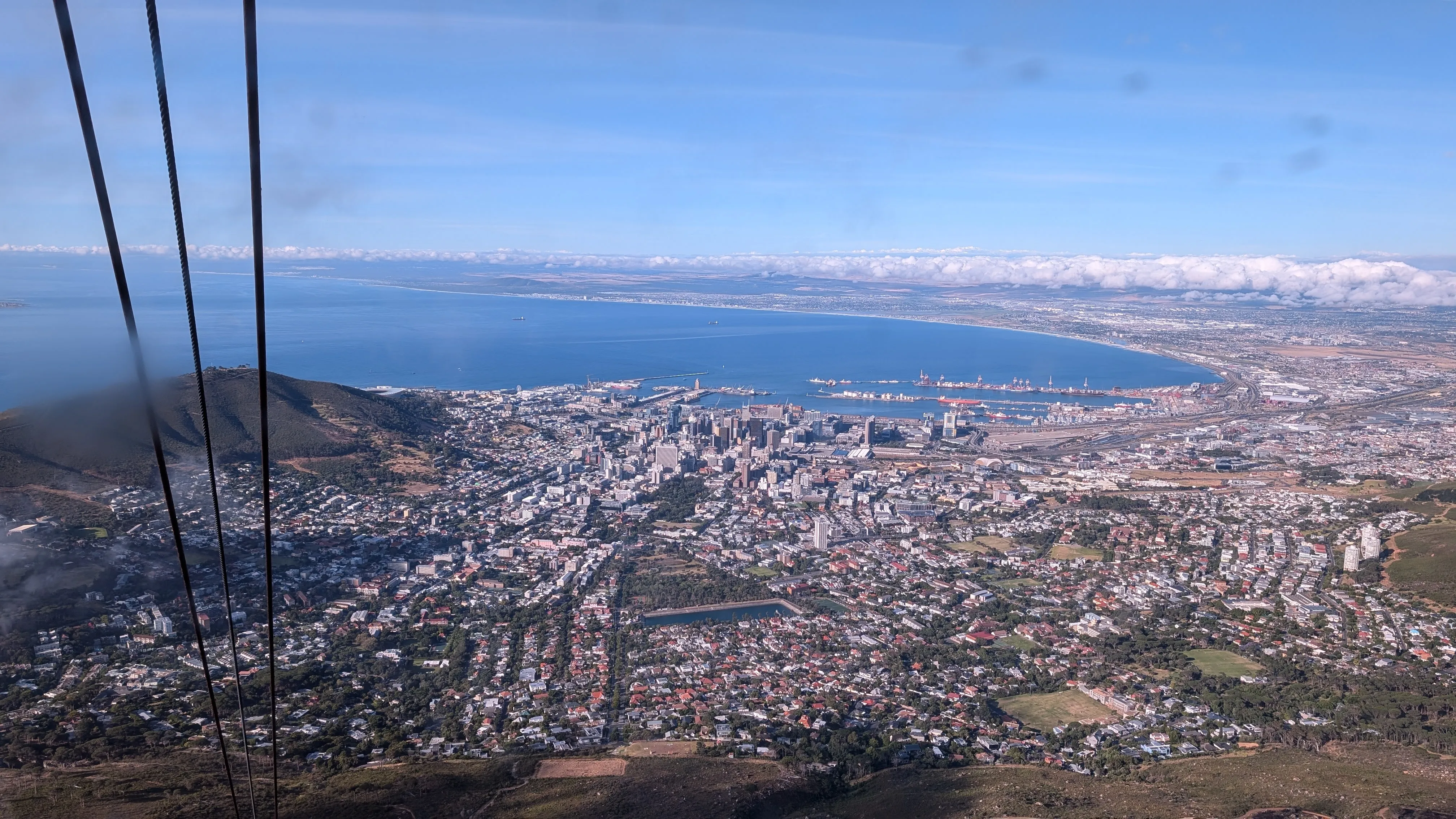 Aerial view of Cape Town showing the city center, coastline, harbor, and surrounding mountains under a clear blue sky.