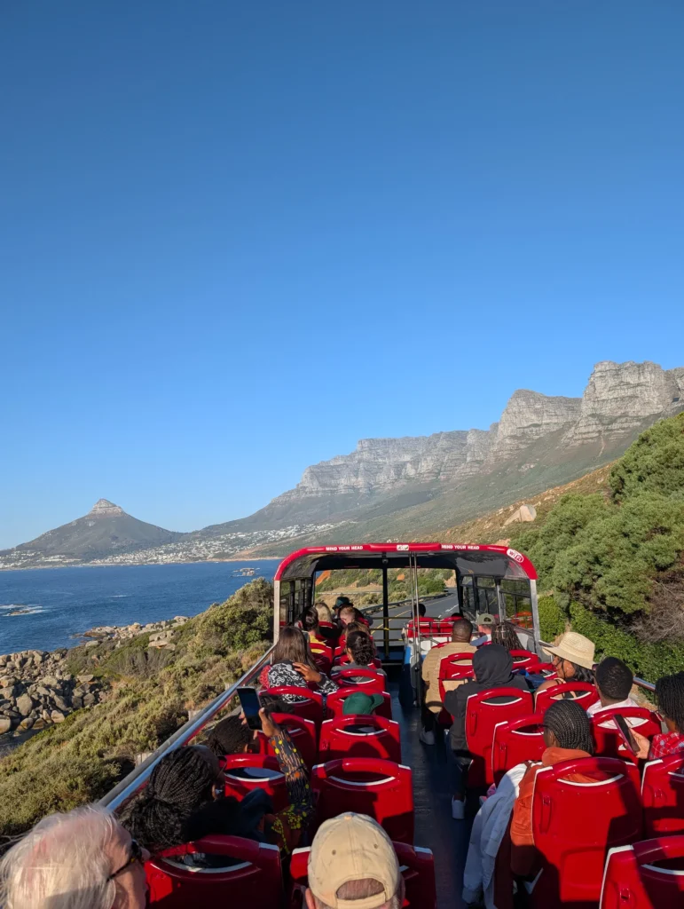 Passengers on an open-top red sightseeing bus driving along Cape Town’s coastal road with Lion’s Head and the Twelve Apostles mountains in the background under a clear blue sky.