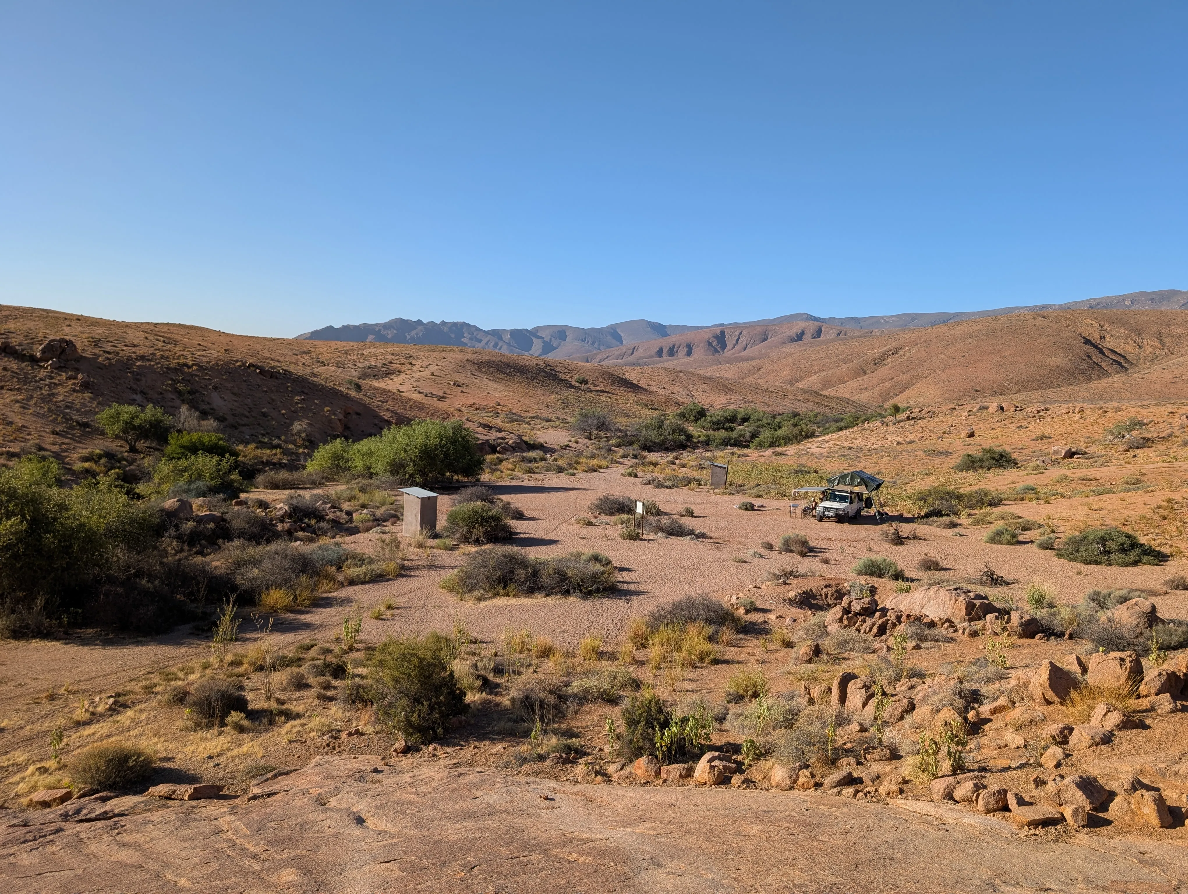 Remote desert campsite set in a wide valley with scattered bushes, rugged hills, and an off-road vehicle parked near a small stone shelter.
