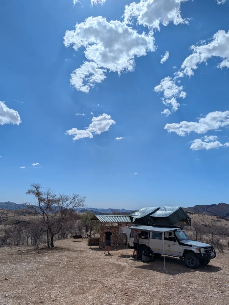 Campsite on a rocky hilltop with a Bushlore Land Cruiser and rooftop tent set up beside a small stone shelter under a bright blue sky with scattered clouds.