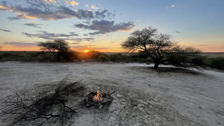 Small campfire burning on a sandy clearing at sunset in Khiding Camp with trees and a colorful sky in the background.