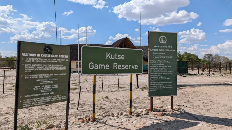 Entrance signs for Khutse Game Reserve standing in sandy terrain under a bright blue sky with scattered clouds.