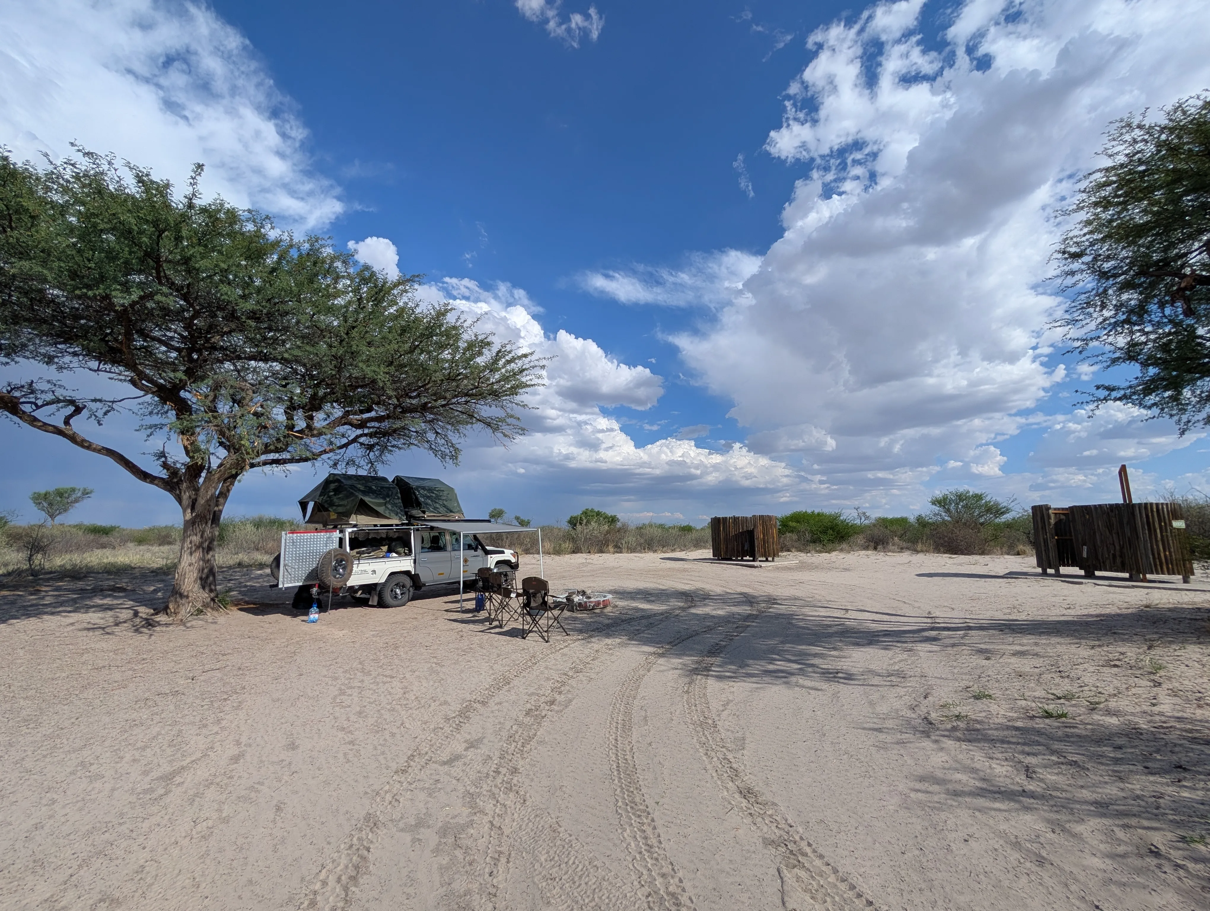 Off-road vehicle with rooftop tent set up under a lone tree at a sandy campsite in Kutse Game Reserve, with wide open skies and scattered clouds above.