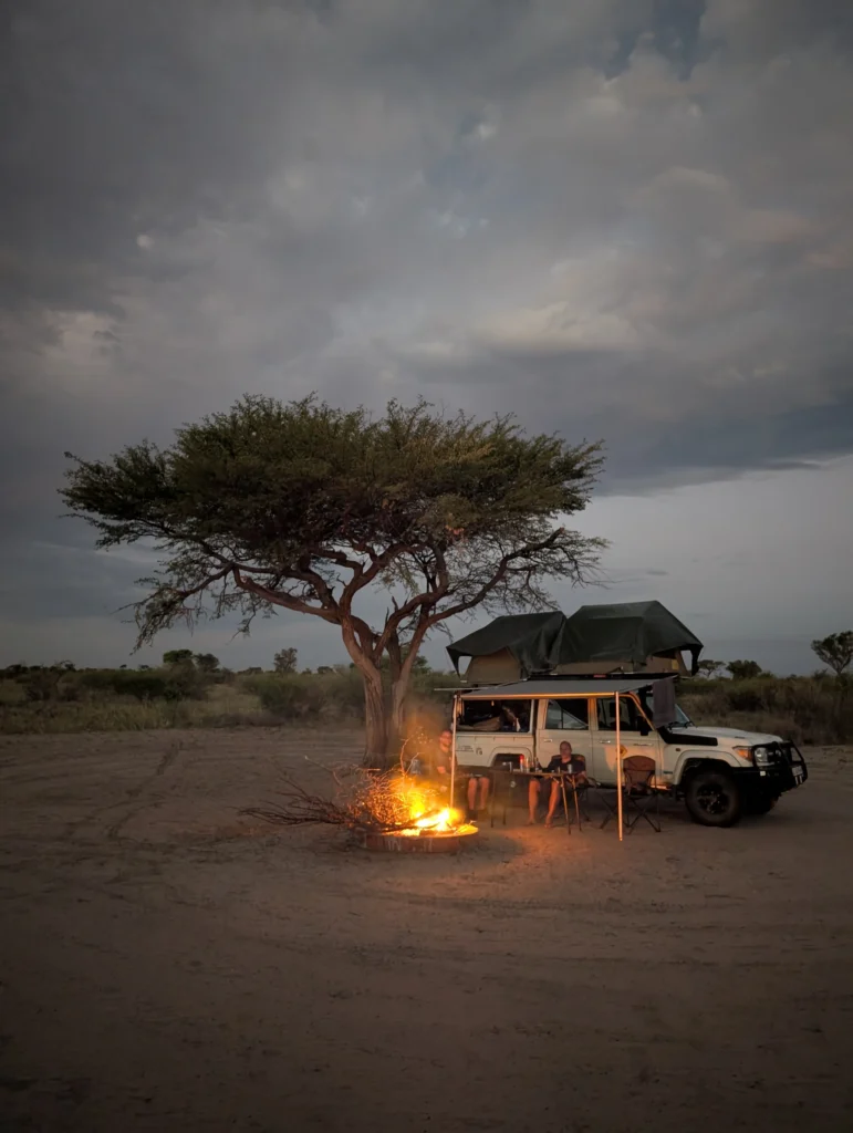 A 4x4 vehicle with a rooftop tent and a campfire at a remote campsite under a cloudy dusk sky.