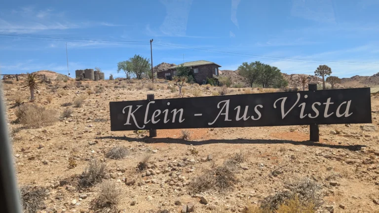 Entrance sign reading “Klein Aus Vista” set in a rocky desert landscape with scattered bushes, trees, and a small stone building in the background.