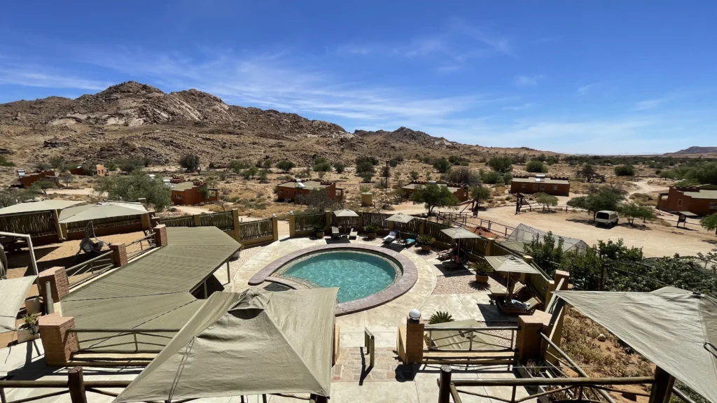 Desert lodge with a circular swimming pool surrounded by shaded seating areas, green canopies, and scattered chalets set against rocky hills under a bright blue sky.