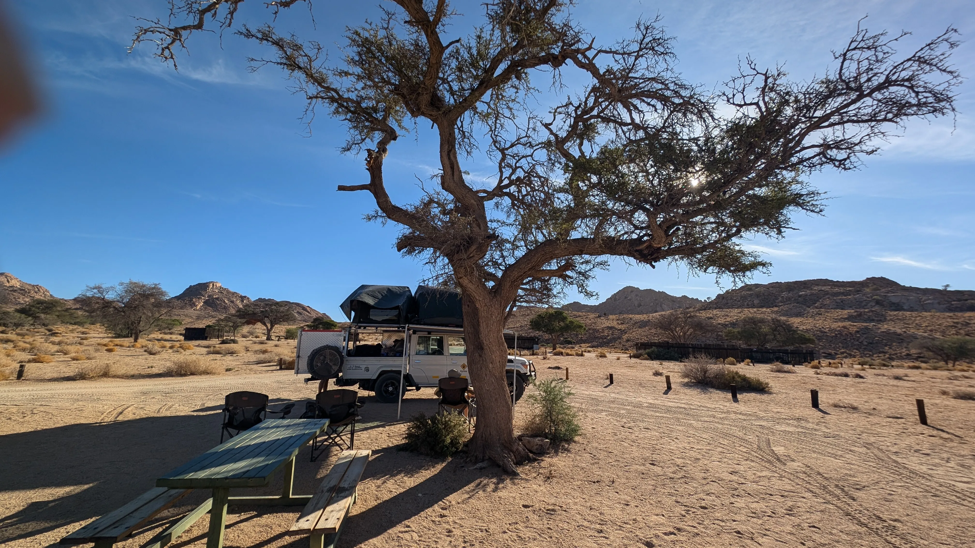 Shaded desert campsite with a rooftop-tent Land Cruiser parked beside a large tree, a picnic table in the foreground, and rocky hills in the background.
