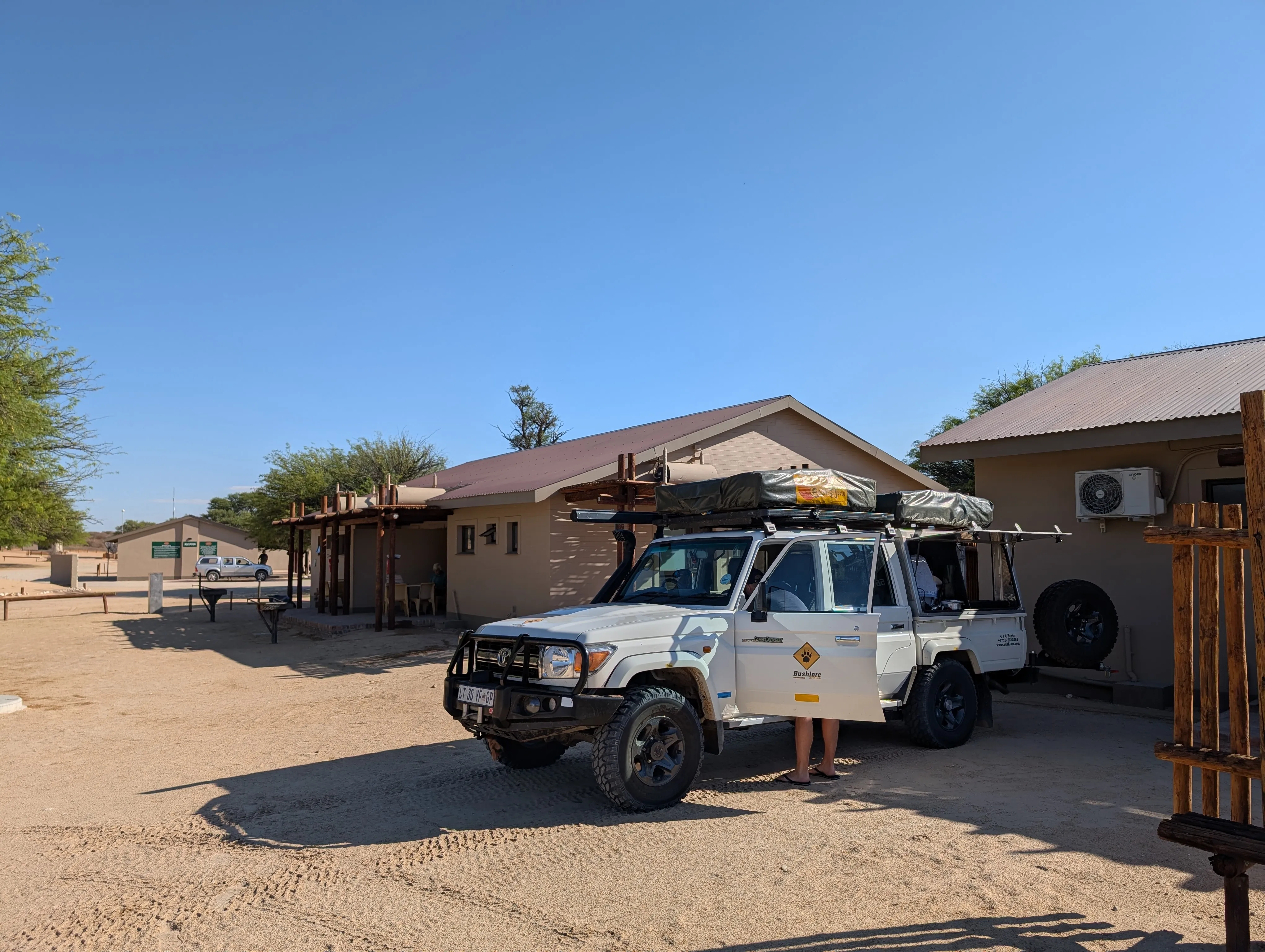A white 4x4 safari vehicle with a rooftop tent parked in the sandy grounds of Nossob Rest Camp.
