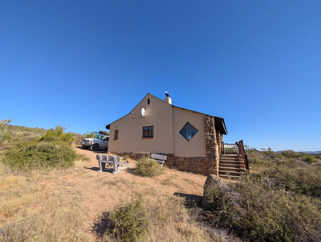 Isolated stone-and-plaster cottage on a hillside with clear blue sky, surrounded by dry shrubland and a parked off-road vehicle nearby.