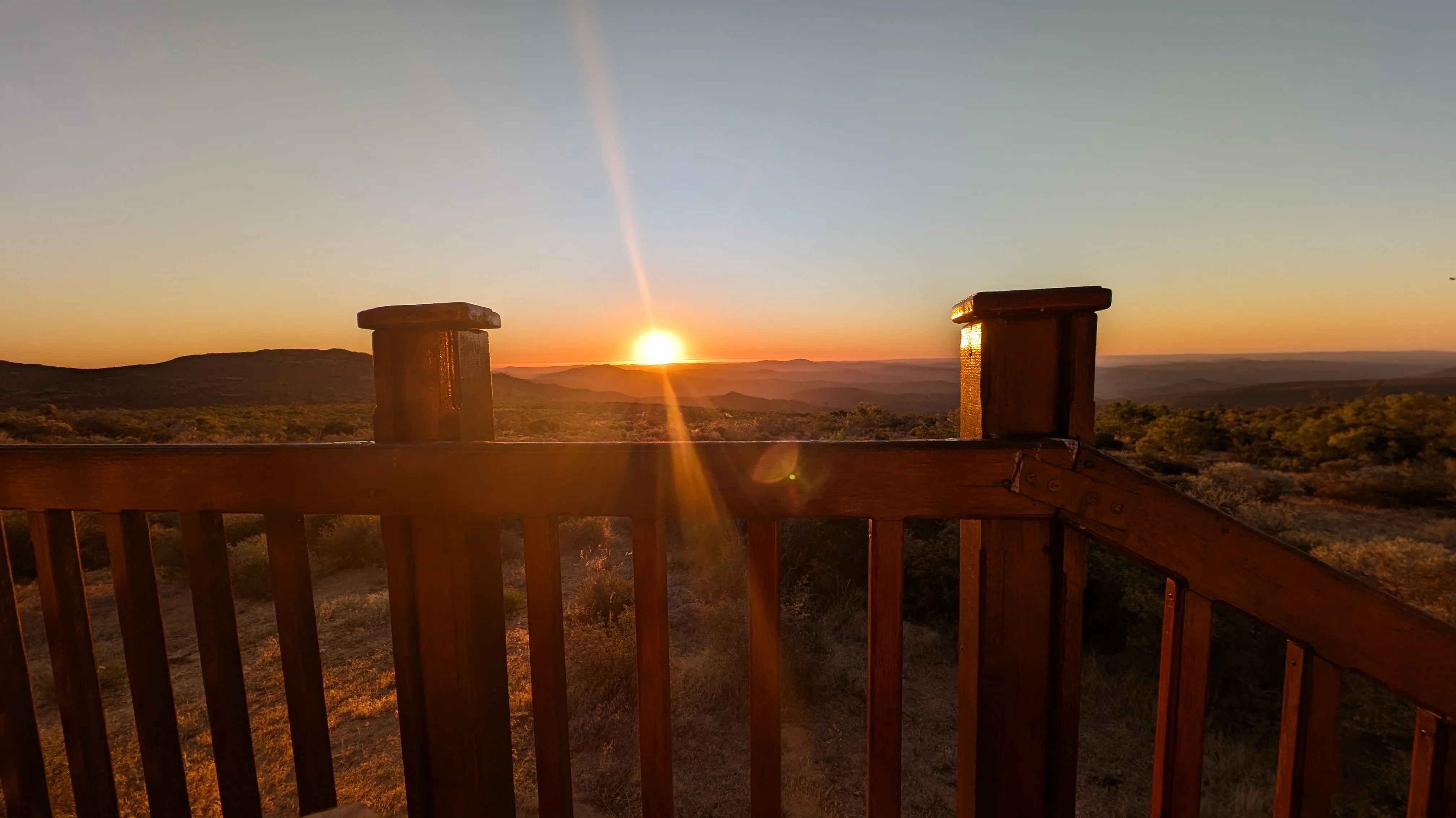 Sunset viewed from a wooden deck overlooking a wide landscape of hills and shrubs, with warm orange light spreading across the horizon.