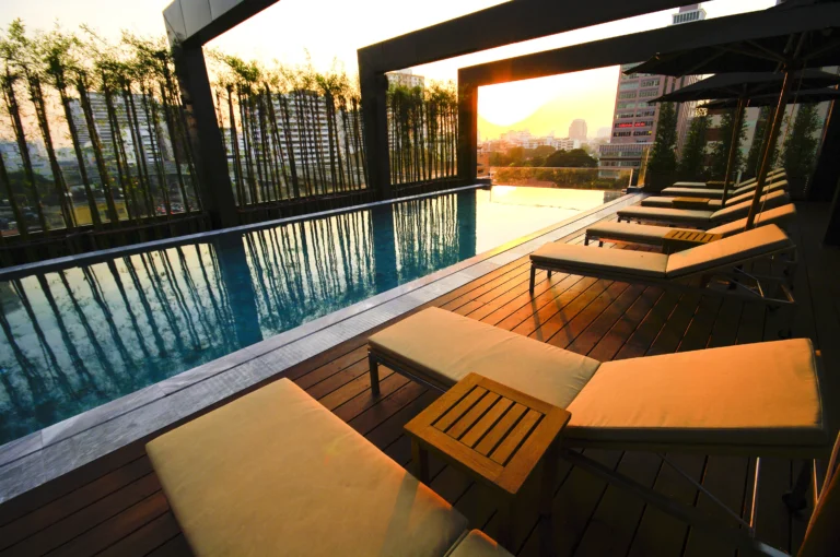 Rooftop pool with beige sun loungers on a wooden deck at sunset, overlooking city buildings and tall bamboo plants in Small Luxury Hotels.