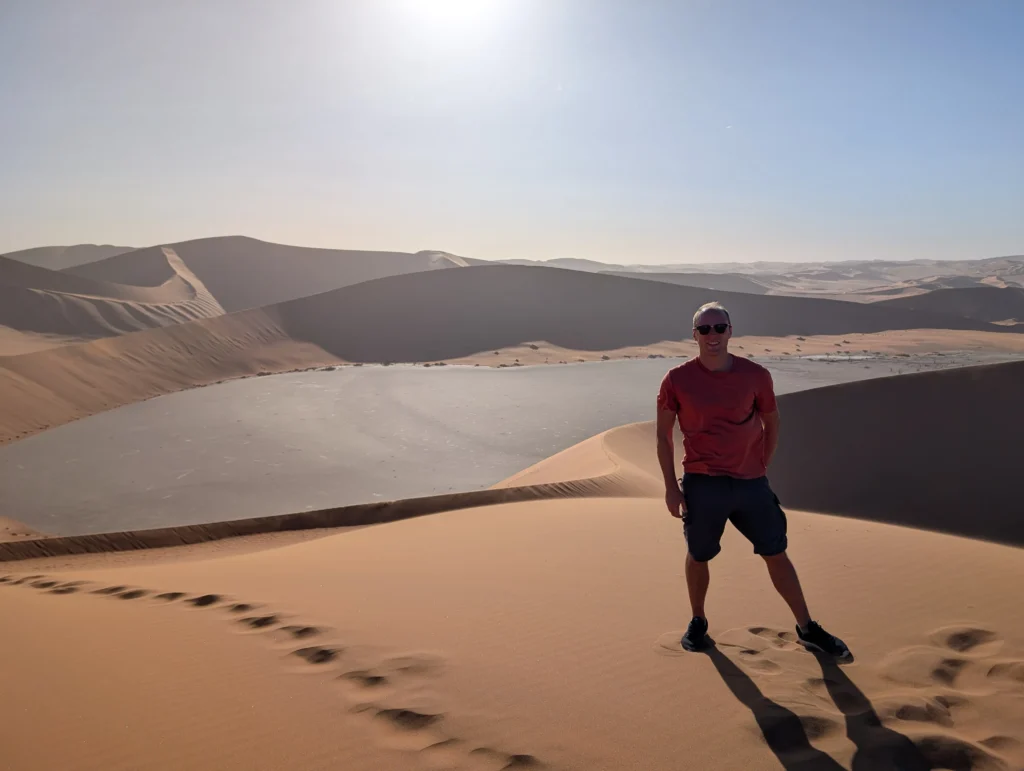 Person standing on a tall sand dune at Sossusvlei in Namibia, overlooking a dry clay pan surrounded by sweeping desert dunes under bright sunlight.