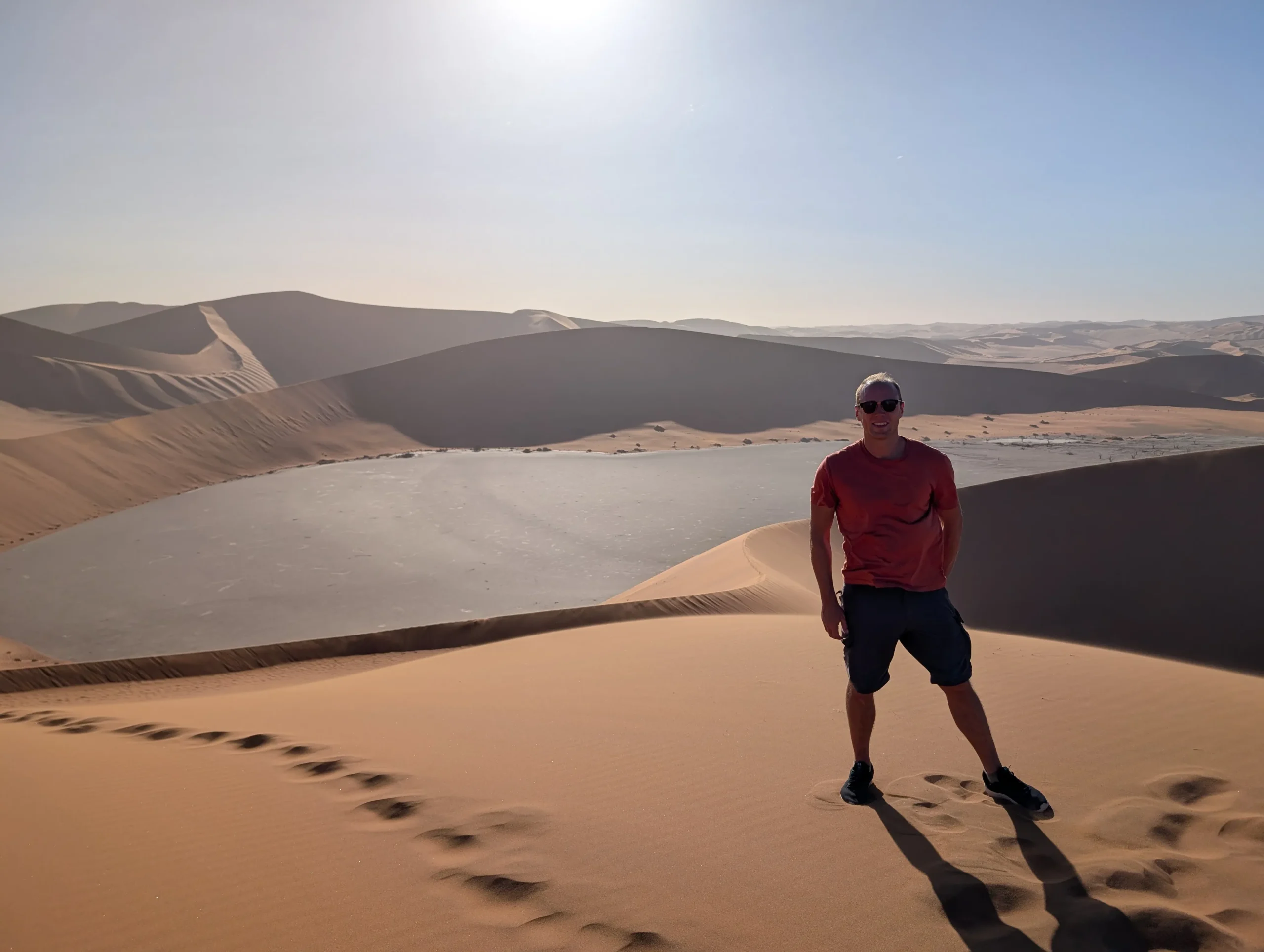 Person standing on a tall sand dune at Sossusvlei in Namibia, overlooking a dry clay pan surrounded by sweeping desert dunes under bright sunlight.
