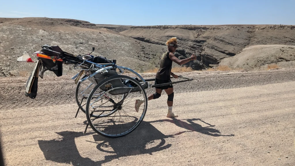 Person running along a gravel road while pulling a large custom cart with gear through a rocky desert canyon landscape.