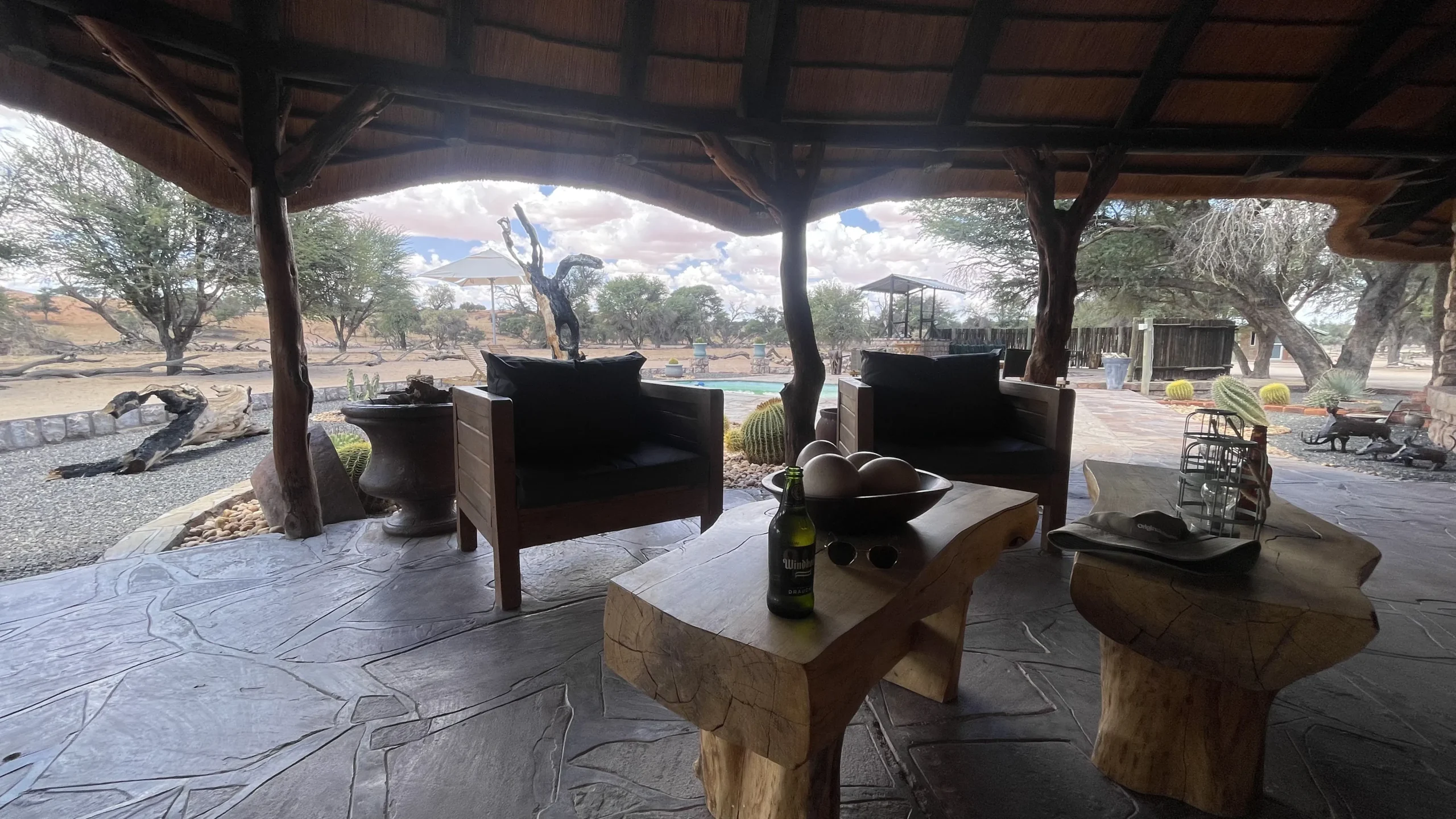 Covered outdoor lodge seating area in Togos Lodge with rustic wooden furniture overlooking a pool and trees in a safari setting.