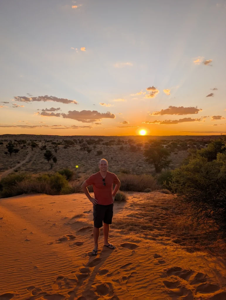 Person standing on a sand dune at sunset with warm orange light, scattered clouds, and a wide desert landscape in the background.