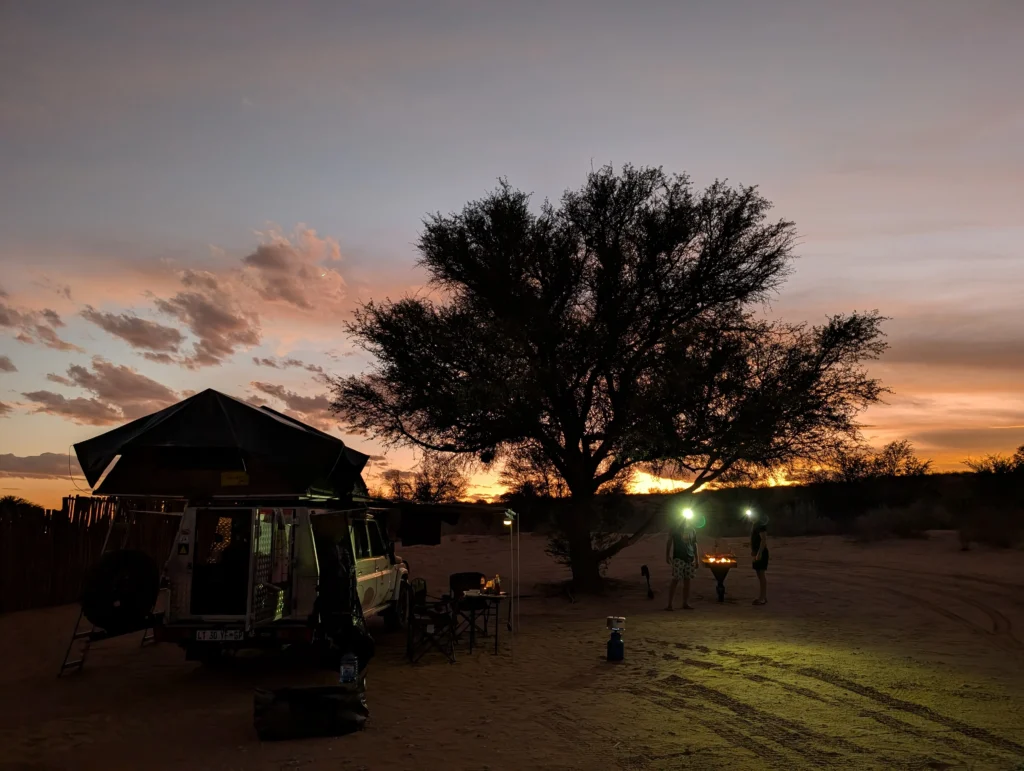 Evening campsite at Twee Rivieren Rest Camp with a rooftop-tent vehicle, chairs, a tree silhouetted against a colorful sunset, and two people preparing a fire with headlamps.