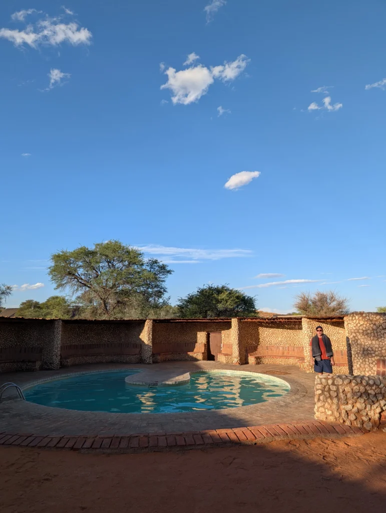 Small circular pool surrounded by brick and stone walls at a desert lodge in Kgalagadi Transfrontier Park, with a person standing nearby under a clear blue sky.