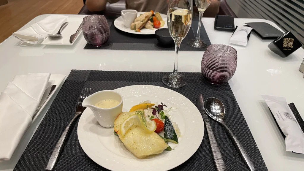 Close-up of fine dining meal with white fish, champagne, and purple glassware in the Qatar Airways lounge.