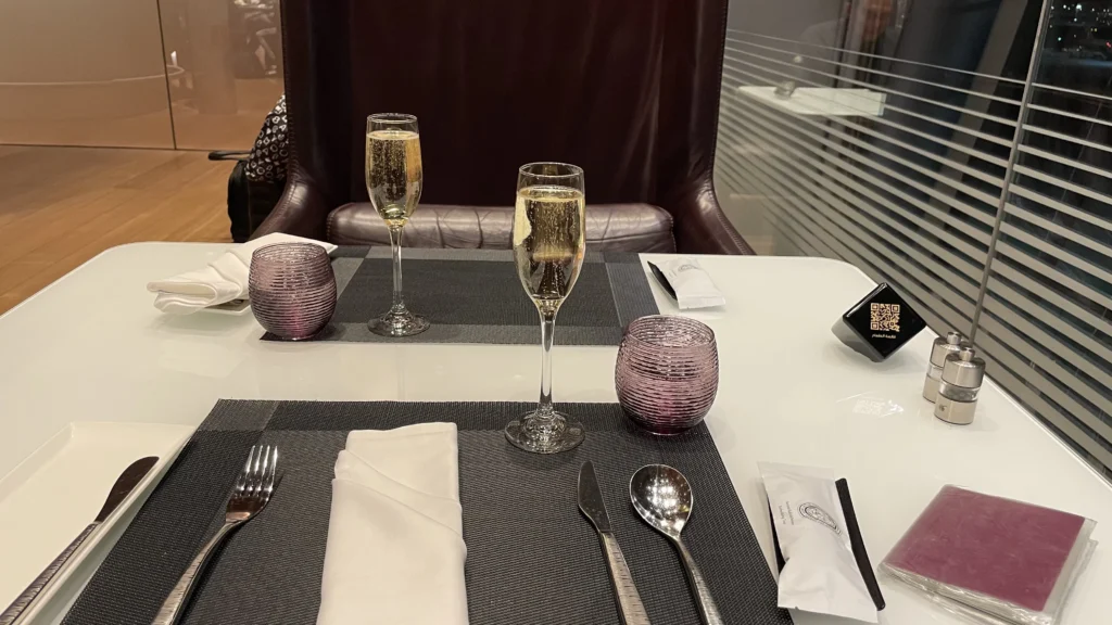 An elegant dining table set for two, likely in an airport lounge, featuring two champagne flutes, purple textured water glasses, silverware, and white napkins arranged on dark placemats, with a large leather high-back chair in the background.