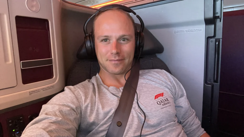 Passenger relaxing in Qatar Airways Business Class Qsuite seat wearing headphones and an F1 Qatar Airways shirt.
