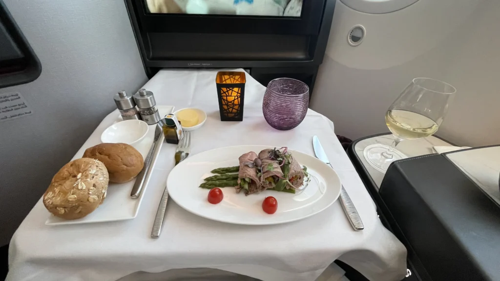 A luxurious business class in-flight meal served on a white tablecloth, featuring an appetizer of roast beef rolls with asparagus and cherry tomatoes, accompanied by fresh bread rolls, a glass of white wine, a textured purple water glass, and a decorative electric candle.