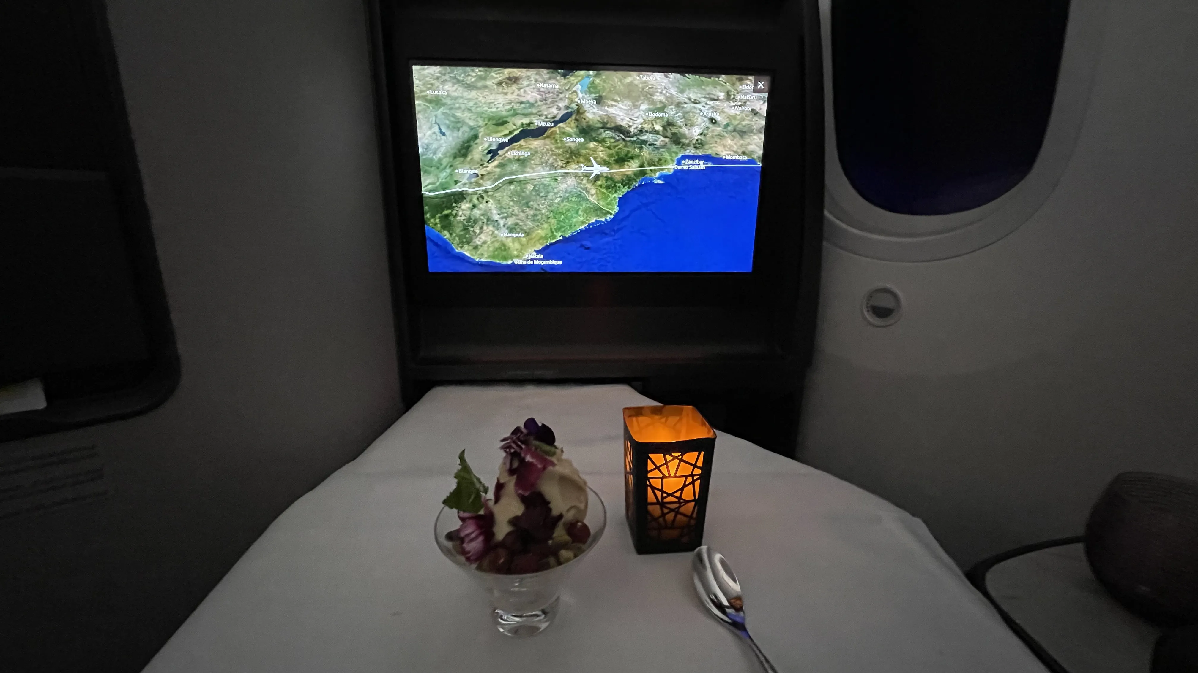 A dessert course served in a dimly lit airplane cabin, featuring a glass bowl of ice cream or mousse with berry toppings next to a glowing electric candle, with a flight map displayed on the entertainment screen in the background.
