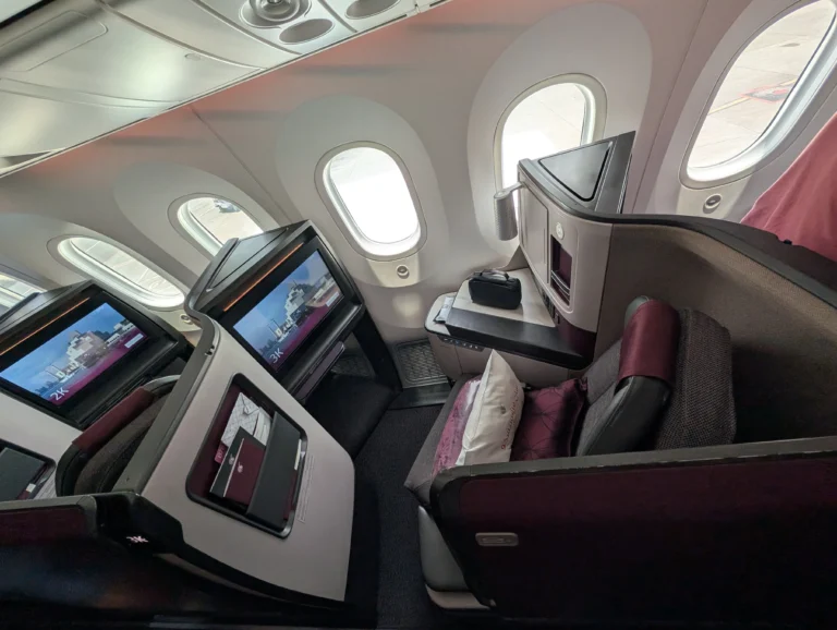 The interior of the qatar business class airplane cabin showing angled seats with maroon and grey upholstery, large windows, and individual entertainment screens.